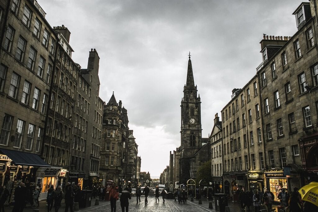 A city street lined with historic stone buildings, pedestrians walking, and a prominent clock tower in the background under a cloudy sky—an inviting scene for those seeking authentic scotland travel.