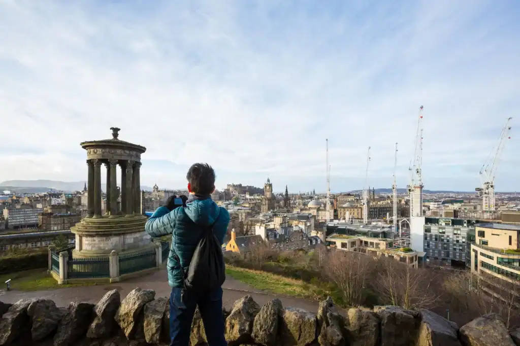 A person in a blue jacket stands on a stone overlook, capturing the cityscape of Edinburgh’s historic buildings and construction cranes—an inspiring moment for those seeking luxury travel Edinburgh experiences under a partly cloudy sky.